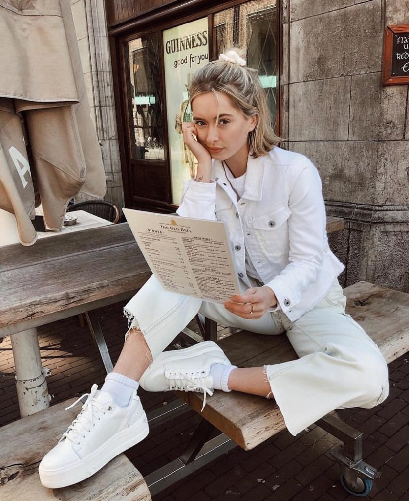 picture of a woman wearing white denim and white sneakers sitting on a bench reading a menu